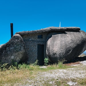 Puede incluir: Una casa de piedra única construida en grandes rocas, con una puerta oscura, chimenea y una pequeña antena. La estructura está ubicada en un paisaje herboso bajo un cielo azul claro, mostrando una mezcla de naturaleza y arquitectura.
