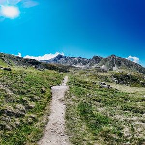 Puede incluir: Un paisaje montañoso con un camino de tierra que conduce a las cumbres. El cielo es de un azul vibrante con un sol brillante. La hierba verde y la vegetación cubren las laderas inferiores, y las montañas cubiertas de nieve están al fondo.