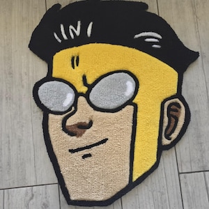 May include: A rug in the shape of a cartoon character's head. The rug features a yellow face, black hair, and gray glasses. The character has a tan nose and mouth, and a black outline. The rug is placed on a wooden floor.