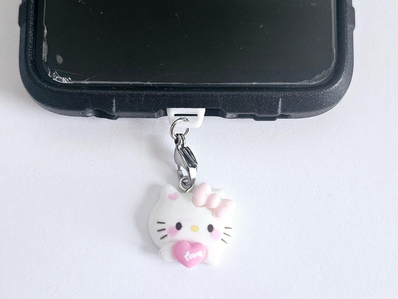 May include: A white Hello Kitty phone charm with a pink bow and a pink heart. The charm is attached to a silver chain and a white clip.