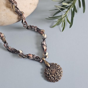 May include: A brown, gold, and black braided necklace with a round, bronze pendant featuring a floral design.