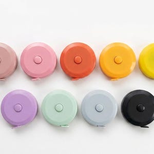 May include: A collection of nine colorful retractable measuring tapes in various shades, including pink, orange, yellow, purple, green, gray, and black. Each tape has a button in the center and a small tab for pulling the tape.