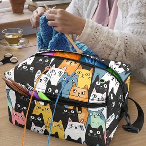 May include: A rectangular yarn storage bag featuring a colorful all-over print of various cartoon cats in black, white, yellow, orange, pink, and blue. The bag has a zippered top, metal grommets for yarn feeding, and a black adjustable shoulder strap.