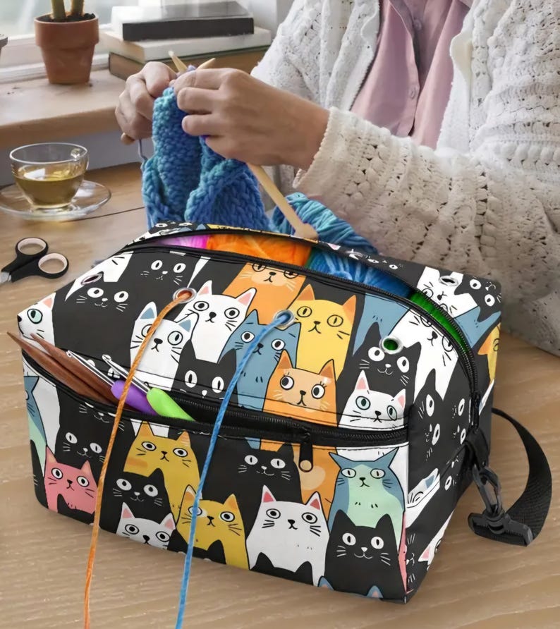 May include: A rectangular yarn storage bag featuring a colorful all-over print of various cartoon cats in black, white, yellow, orange, pink, and blue. The bag has a zippered top, metal grommets for yarn feeding, and a black adjustable shoulder strap.