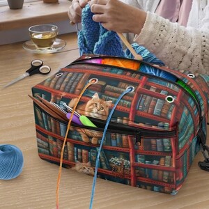 May include: A rectangular yarn storage bag featuring a printed design of a library bookshelf with a ginger cat. The bag has a zippered top, metal grommets for yarn strands, and a black shoulder strap. Several crochet hooks are tucked into the front pocket.