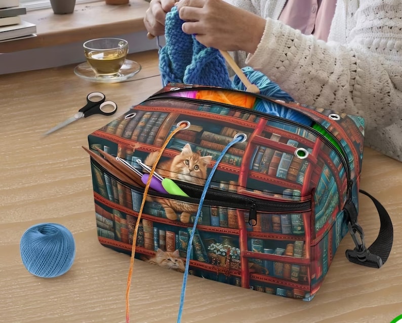 May include: A rectangular yarn storage bag featuring a printed design of a library bookshelf with a ginger cat. The bag has a zippered top, metal grommets for yarn strands, and a black shoulder strap. Several crochet hooks are tucked into the front pocket.