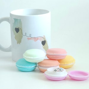 May include: A white ceramic mug with a watercolor design of two states connected by a line with hearts at the ends. The mug is surrounded by six pastel-colored macaron-shaped boxes, one of which is open and reveals a small silver ring with a flower design.