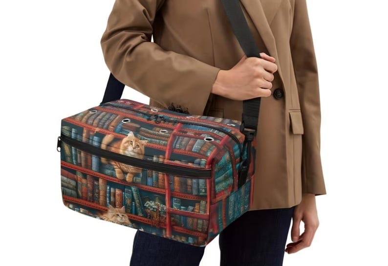 May include: A rectangular pet carrier bag featuring a vibrant print of a library bookshelf filled with numerous books and two orange tabby cats. The bag includes a black adjustable shoulder strap, a zippered top closure, and silver metal grommets.