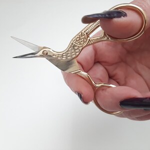 May include: Gold-coloured bird-shaped scissors with intricate details. The scissors are held in a hand with dark nail polish.