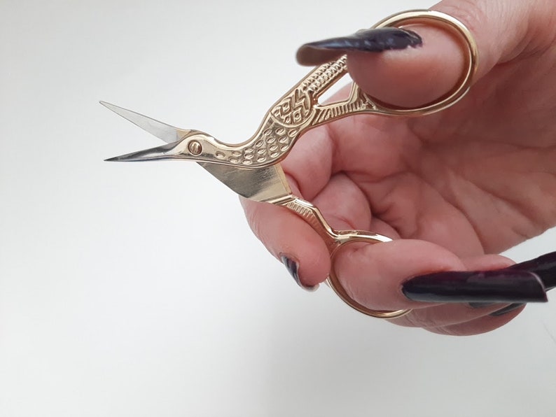 May include: Gold-coloured bird-shaped scissors with intricate details. The scissors are held in a hand with dark nail polish.