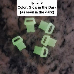 May include: Five glow-in-the-dark plastic clips, shaped like a small person with a rounded head and arms, and a rectangular body. The clips are arranged in a cluster on a dark surface.