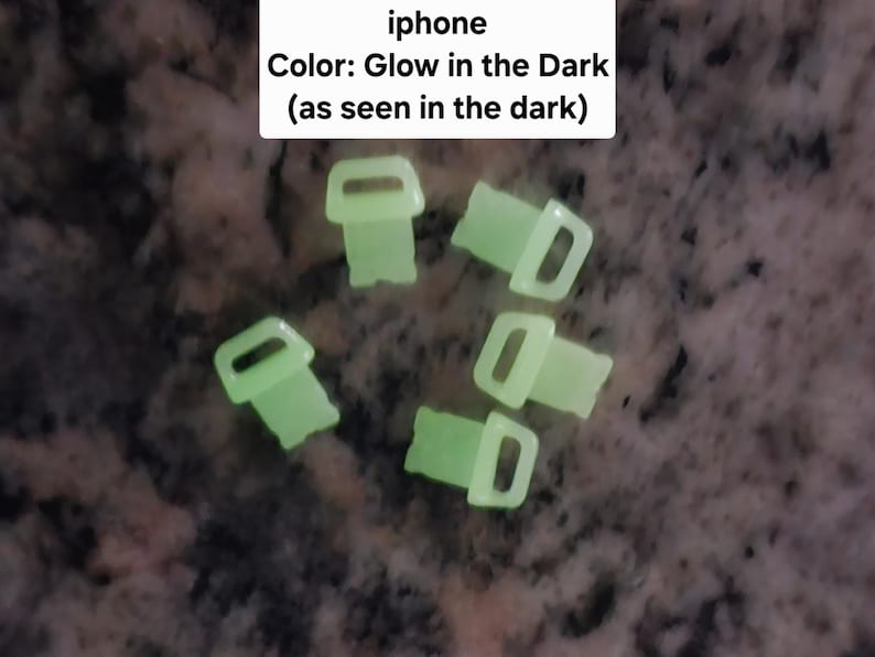 May include: Five glow-in-the-dark plastic clips, shaped like a small person with a rounded head and arms, and a rectangular body. The clips are arranged in a cluster on a dark surface.