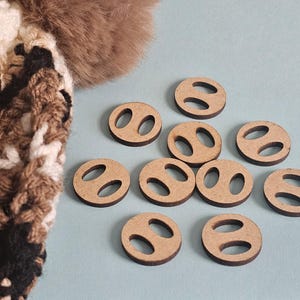 May include: A collection of ten small, round wooden buttons with two oval holes each, arranged on a light blue surface. A portion of a brown and white knitted item with a fur accent is visible in the upper left corner.