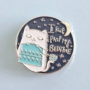 May include: A white cat with a teal knitted scarf and yarn ball sits on a black background with stars and a crescent moon. The text "I knit past my bedtime" is written in gold lettering.