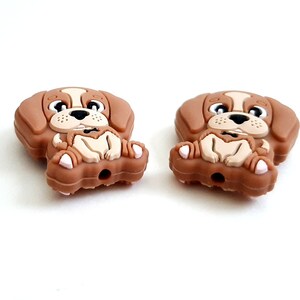Puede incluir: Dos abalorios de perro de silicona marr&oacute;n con un dise&ntilde;o de beagle de dibujos animados. Los perros est&aacute;n sentados con las patas cruzadas y tienen una expresi&oacute;n feliz en sus caras.