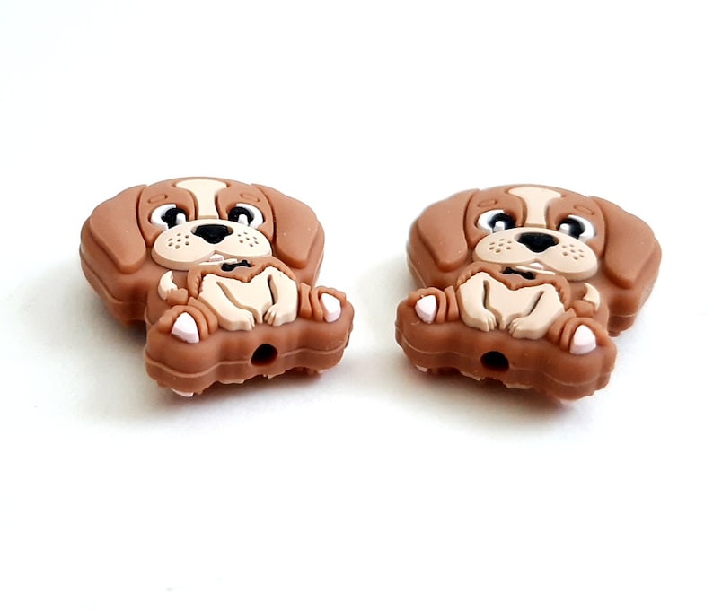 Puede incluir: Dos abalorios de perro de silicona marr&oacute;n con un dise&ntilde;o de beagle de dibujos animados. Los perros est&aacute;n sentados con las patas cruzadas y tienen una expresi&oacute;n feliz en sus caras.