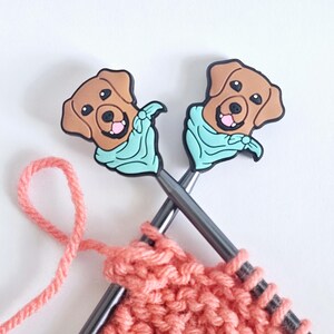 Puede incluir: Dos agujas de tejer con adornos de cabeza de perro marr&oacute;n que llevan bandanas azules. Las agujas est&aacute;n cruzadas y tienen un hilo de color coral sujeto.