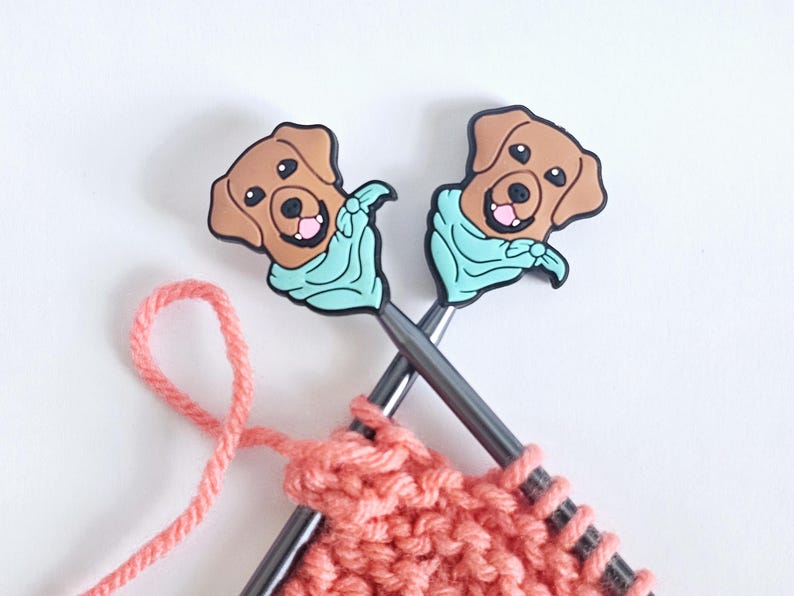 Puede incluir: Dos agujas de tejer con adornos de cabeza de perro marr&oacute;n que llevan bandanas azules. Las agujas est&aacute;n cruzadas y tienen un hilo de color coral sujeto.