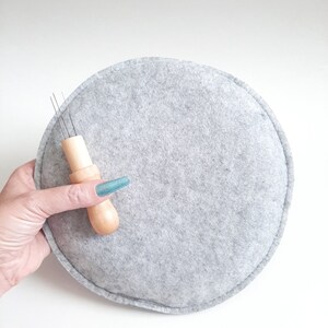 Op de afbeelding: Een grijze viltcirkel met een houten naaldgereedschap met drie naalden. Het gereedschap wordt vastgehouden door een hand met blauwe nagellak.