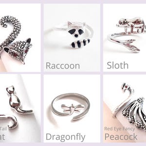 May include: A collection of silver-toned animal-themed rings. Designs include a fox, raccoon, sloth, cat, dragonfly, and peacock. Each ring is shown on a white surface with the animal's name below it. The peacock ring has a red gem for an eye.