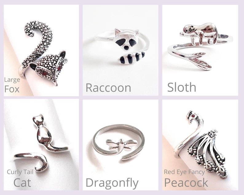May include: A collection of silver-toned animal-themed rings. Designs include a fox, raccoon, sloth, cat, dragonfly, and peacock. Each ring is shown on a white surface with the animal's name below it. The peacock ring has a red gem for an eye.