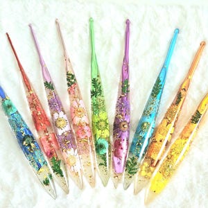 May include: A collection of colorful crochet hooks, each encased in clear resin and adorned with dried flowers and gold flakes. The hooks feature a variety of colors, including blue, green, purple, and yellow.