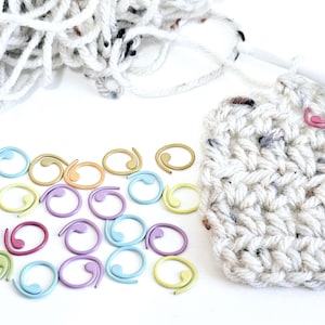 May include: A close-up of a white crocheted square with a crochet hook and a pile of white yarn. There are also a variety of colourful stitch markers in the shape of a circle with a tail.