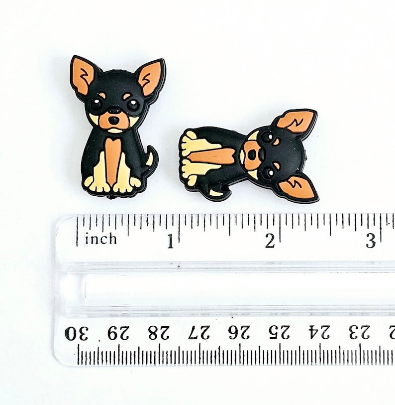 Puede incluir: Dos abalorios de perro chihuahua negro y marr&oacute;n, de aproximadamente 2,5 cm de tama&ntilde;o, sobre una regla.