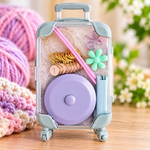 May include: A light blue miniature suitcase-shaped storage case containing knitting supplies, including pink knitting needles, gold-toned stitch markers with the number one, a light purple circular tape measure, and a light green flower-shaped tool.