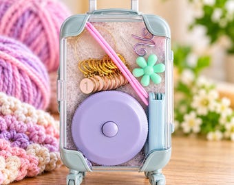 May include: A light blue miniature suitcase-shaped storage case containing knitting supplies, including pink knitting needles, gold-toned stitch markers with the number one, a light purple circular tape measure, and a light green flower-shaped tool.