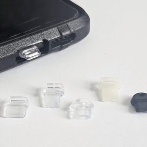 May include: A close-up of five small plastic phone port covers. Three are clear, one is white, and one is black.