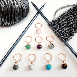May include: A collection of gemstone stitch markers with copper wire loops, arranged with knitting needles, yarn, and a knitted swatch. The gemstones are various colors, including pink, green, purple, gray, white, blue, and brown. The yarn is a gray and silver blend.