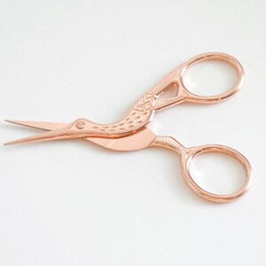 May include: Rose gold scissors with a bird design. The scissors have a small, pointed blade and a larger, rounded blade.