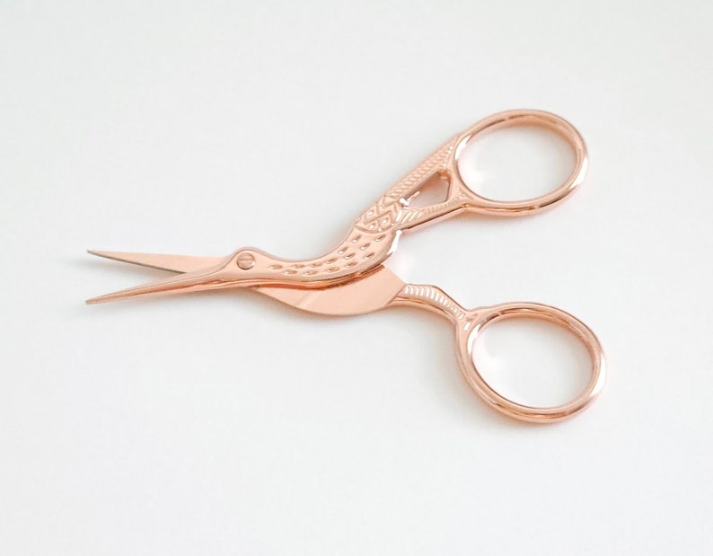 May include: Rose gold scissors with a bird design. The scissors have a small, pointed blade and a larger, rounded blade.