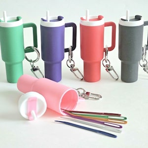 May include: A collection of colorful, miniature tumbler-shaped keychains in green, purple, red, and gray, each with a white lid and handle. A pink container holds several colored straws and a white lid.