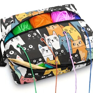 May include: A rectangular yarn storage bag featuring a repeating pattern of cartoon cats in black, white, orange, and blue. The bag has a zippered top and front pocket with metal grommets for feeding yarn strands through, and a black shoulder strap.