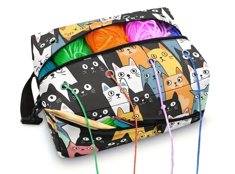 May include: A rectangular yarn storage bag featuring a repeating pattern of cartoon cats in black, white, orange, and blue. The bag has a zippered top and front pocket with metal grommets for feeding yarn strands through, and a black shoulder strap.