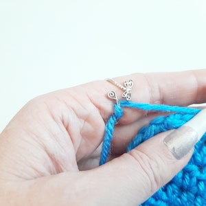 May include: Close-up of a hand crocheting with bright blue yarn. A silver dragonfly ring is on the index finger, guiding the yarn. A green crochet hook is visible, along with the partially completed blue crochet project.