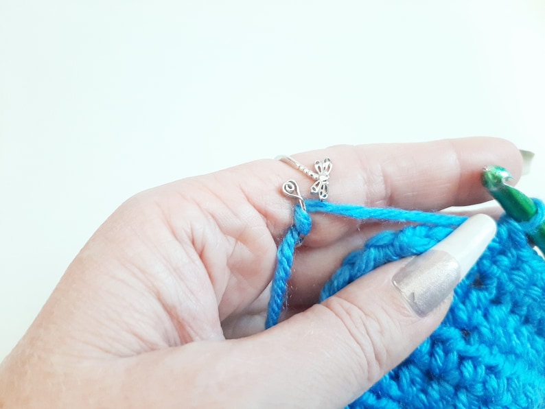May include: Close-up of a hand crocheting with bright blue yarn. A silver dragonfly ring is on the index finger, guiding the yarn. A green crochet hook is visible, along with the partially completed blue crochet project.