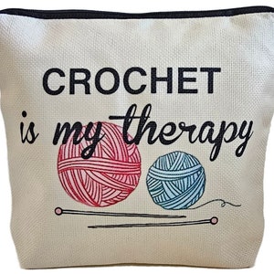 May include: A white canvas zippered pouch with the text "CROCHET is my therapy" printed in black. Two balls of yarn, one pink and one blue, are illustrated with knitting needles.