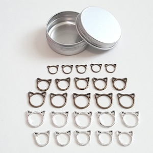 May include: A set of 20 silver and bronze cat-shaped stitch markers in a silver tin. The markers are arranged in two rows, with the silver markers on the bottom and the bronze markers on the top.