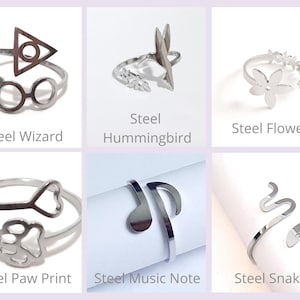 May include: A collection of silver-colored steel rings with various designs. The rings include a wizard-themed ring, a hummingbird, flower, paw print, music note, and snake. Each ring has the design name printed below it.