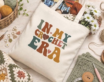 May include: A cream-colored canvas tote bag featuring the text "IN MY CROCHET ERA" in a multi-colored, retro-style font. The design includes small heart accents and a heart-shaped graphic replacing the letter O in the word crochet.