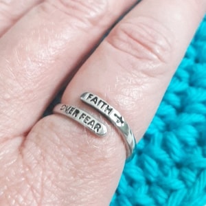 May include: A silver ring with the words "Faith Over Fear" engraved on it. The ring is worn on a finger and the words are visible.