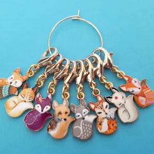 May include: Set of eight gold-tone fox stitch markers with colorful enamel designs. Each marker features a different fox design with a variety of colors and patterns.
