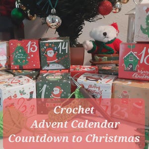 May include: A collection of square cardboard boxes featuring numbered Christmas-themed illustrations, including a green dinosaur, Santa Claus, and reindeer. A ball of green yarn and a crochet hook are positioned in front of the boxes.