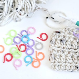 May include: A selection of colourful stitch markers in various shades, such as red, purple, and green, beside a crocheted swatch and a ball of white yarn. A crochet hook is also visible.