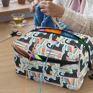 May include: A rectangular yarn storage bag featuring a light blue background with a repeating pattern of black cats, colorful books, and potted plants. The bag has a black zippered top, a front pocket with yarn guide grommets, and a black strap.