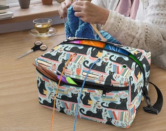 May include: A rectangular yarn storage bag featuring a light blue background with a repeating pattern of black cats, colorful books, and potted plants. The bag has a black zippered top, a front pocket with yarn guide grommets, and a black strap.