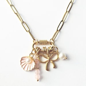 May include: Gold-coloured necklace with a chain link design. The necklace features a seashell charm, a bow charm, and a pearl charm. The seashell is pink and white. The necklace also has pink beads.